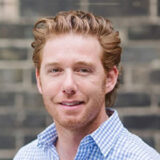I don’t know who this is, but the image shows a person with short, wavy reddish hair, wearing a light blue checked shirt, smiling slightly in front of a blurred brick background.