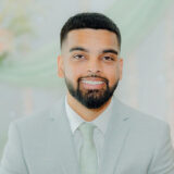 A smiling man wearing a light gray suit and tie stands against a soft, blurred background, exuding a sense of warmth and confidence.