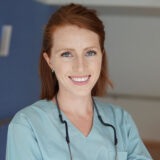 A smiling person with red hair and blue scrubs stands in a medical or dental setting, wearing a stethoscope around their neck.