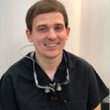 A person in a dental uniform is smiling at the camera, wearing dental loupes around their neck.