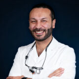 A smiling dentist in a white lab coat, arms crossed, with a dental loupe around his neck, standing against a dark background.