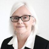 A smiling woman with white hair and glasses wearing a black blazer and white shirt against a plain background.