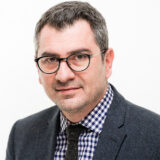 Man with glasses wearing a checkered shirt and dark blazer against a plain white background.