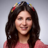 Smiling woman with long dark hair, wearing a red patterned top and a colorful beaded headband, in front of a neutral background.