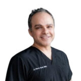 A man wearing a black medical scrub top smiles at the camera against a white background.