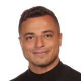 A person with short dark hair smiling, wearing a black shirt, against a white background.