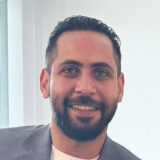 Smiling person with dark hair and a beard, wearing a blazer.