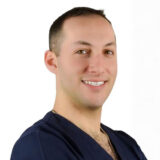 Person smiling warmly, wearing a navy blue dental scrubs.