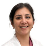 A smiling woman with dark hair, wearing a white coat, possibly indicating a professional in a healthcare setting, against a plain white background.