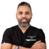 A dentist with a friendly smile, wearing a black scrub top, arms crossed, set against a plain white background.