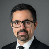 A man with short dark hair, glasses, and a beard, dressed in a suit and tie, poses against a gray background.