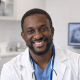 A smiling dentist in a white coat, wearing a stethoscope, sits in a dental office with medical equipment and a monitor in the background.