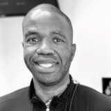 A person with a friendly smile, wearing a dark jacket, poses for a photo in a well-lit room.