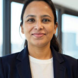 A person in a suit is smiling in an indoor setting with large windows in the background.