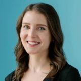 A woman with long brown hair and a friendly smile, wearing a black top, is posed against a light blue background.