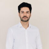 A man with short dark hair and a beard wearing a white button-up shirt, standing against a plain background.
