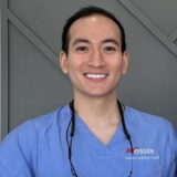 Smiling person wearing blue dental scrubs with a stethoscope around their neck, standing indoors.