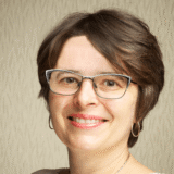 A person with short brown hair and glasses, smiling warmly.
