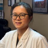 A dentist wearing glasses and a white coat, smiling in an office setting with framed pictures on the wall.
