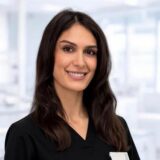 A person with long dark hair smiles warmly, wearing black professional attire in a bright, modern office environment.