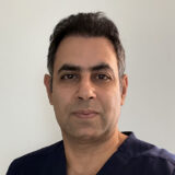 A person with short, dark hair wearing a dark blue shirt, standing against a plain light background.