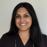 A person with long dark hair smiles at the camera against a plain light background.