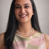 A woman with long dark hair smiles warmly, wearing a floral dress and a pearl necklace against a neutral background.