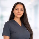 A professional woman with long dark hair smiles gently, wearing a dark medical scrub top, standing in a bright, softly focused environment.