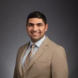 A man in a beige suit and striped tie smiles against a dark gray background.