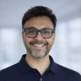 A person with glasses and a beard smiles warmly against a blurred background, wearing a dark polo shirt.