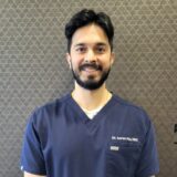 A person with dark hair and a beard is smiling warmly, wearing navy blue dental scrubs against a patterned background.