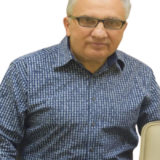 A smiling dentist in glasses and a checkered shirt sits beside a dental chair, ready to assist patients.