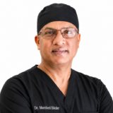A person wearing black medical scrubs and a cap stands with arms crossed. They are wearing glasses and looking directly at the camera.