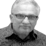 An older person with glasses and short hair wearing a checkered shirt, looking at the camera with a neutral expression.