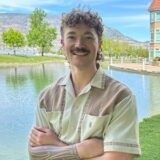 A smiling person with curly hair and a mustache stands by a serene lake with trees and a building in the background.