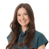 A woman with long brown hair smiles warmly at the camera, wearing a teal blouse.