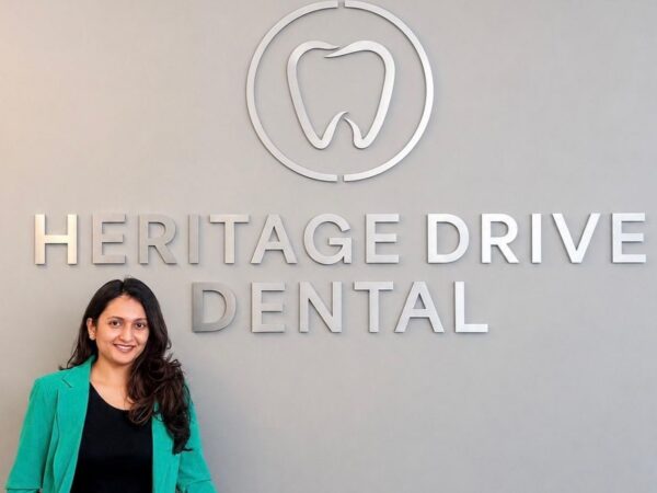 A woman in a green jacket stands smiling in front of a wall sign that reads "Heritage Drive Dental," featuring a tooth logo above the text.