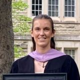 A graduate in a black gown and purple sash holds a framed certificate while standing outdoors in front of a stone building.