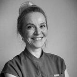 A smiling woman with light hair in a bun, wearing a dental uniform, looking friendly and professional.