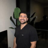 A person wearing a black shirt smiling warmly, standing indoors next to a green plant.