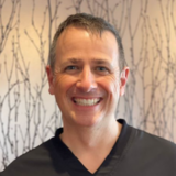 A smiling person in a dental office setting with a patterned background wall.