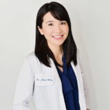 A smiling dentist wearing a white coat and dark blue shirt stands against a plain background.