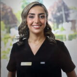 A smiling dental professional in a black uniform stands outside, with trees and a sunny background.