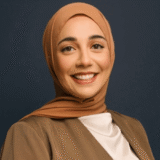 A person wearing a brown hijab and white shirt, smiling against a dark background.