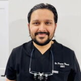 A smiling dentist in a black uniform with magnifying glasses around his neck stands against a light background.