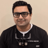 A dentist wearing glasses and a dental uniform with a calm expression, ready to assist patients.