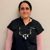 A dentist in scrubs smiles warmly at the camera, wearing magnifying loupes around her neck.