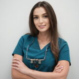 A dentist in teal scrubs smiles confidently with arms crossed, wearing dental loupes around her neck.