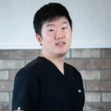 A friendly individual in a black scrub top stands in front of a brick and white wall, smiling gently.