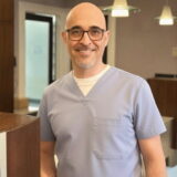 A smiling dentist in light blue scrubs stands in a warmly lit dental office, conveying a welcoming and professional atmosphere.